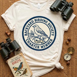 May include: Cream-colored t-shirt with a circular design featuring a bird on a branch, text reading "Backyard Birding Society Charter Member Est." Binoculars and a vintage bird guide are also visible.