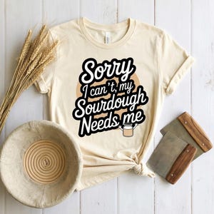 Puede incluir: Una camiseta color crema con el texto "Sorry I can't, my Sourdough Needs me" en un diseño en blanco y negro. La camiseta está anudada en la parte inferior. También se ven una cesta de pan, espigas de trigo y raspadores de masa.