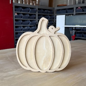 May include: A light-colored wooden cutout in the shape of a pumpkin with a small duck silhouette attached to the stem. The object features a layered, dimensional design with visible wood grain and smooth, precision-cut edges throughout the piece.