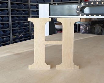 May include: A freestanding capital letter H cut from light-colored wood with a visible grain pattern. The letter features a classic serif typeface design with straight vertical sides and a horizontal crossbar, standing upright on a flat surface.