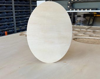 May include: A smooth, light-colored oval wooden plaque with a natural grain pattern. The piece is unfinished, featuring a clean, flat surface and rounded edges, standing upright as a blank canvas for crafting, painting, or decorative projects.