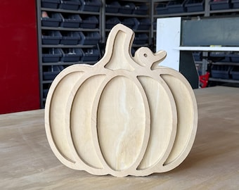 May include: A light-colored wooden cutout in the shape of a pumpkin with a small duck silhouette attached to the stem. The object features a layered, hollowed-out design with visible wood grain and smooth, sanded edges throughout the piece.