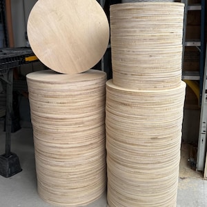 May include: Stacks of unfinished, round wooden discs. One large disc rests atop a stack, with two other stacks of varying heights. The wood has a light, natural color, and the edges of the discs are visible.