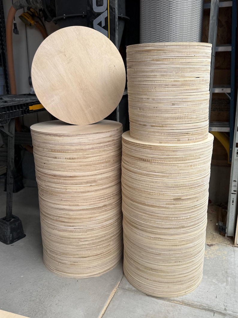 May include: Stacks of unfinished, round wooden discs. One large disc rests atop a stack, with two other stacks of varying heights. The wood has a light, natural color, and the edges of the discs are visible.