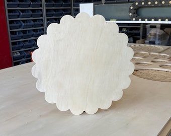 May include: A circular wooden plaque with a scalloped edge. The object is made of light-colored, unfinished plywood with a visible wood grain pattern. The surface is flat and smooth, featuring a consistent, natural beige tone throughout the entire piece.
