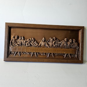 May include: A brown wooden relief carving of the Last Supper, depicting Jesus and his disciples seated at a table. The artwork is framed in a dark brown wooden frame. The figures are carved in detail, with visible textures and shadows.