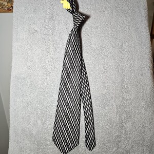 May include: A black and white patterned tie with a repeating oval design. The tie is knotted at the top and hangs vertically. The background is a light gray, textured surface.