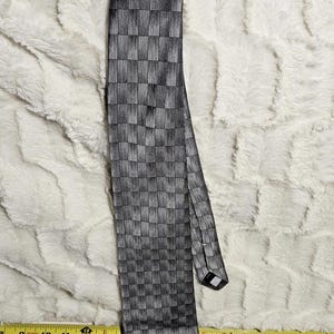 Könnte beinhalten: Eine schwarz-graue Krawatte mit geometrischem Muster. Die Krawatte ist oben geknotet und liegt flach, wodurch ihr Design zur Geltung kommt. Das Muster besteht aus kleinen, vertikalen und horizontalen Linien, die einen karierten Effekt erzeugen. Die Krawatte ist etwa 147 cm lang.