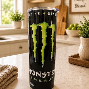 K&ouml;nnte beinhalten: Ein hoher, zylindrischer schwarzer Becher mit dem leuchtend gr&uuml;nen Monster Energy Krallen-Logo auf der Vorderseite. Der obere Teil des Bechers zeigt den Text TAURINE + GINSENG, w&auml;hrend unten das Monster Energy Drink Logo zu sehen ist.