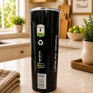 K&ouml;nnte beinhalten: Ein schwarzer zylindrischer Becher mit transparentem Deckel, der ein gr&uuml;n-wei&szlig;es Energy 100%-Symbol, ein Recycling-Symbol, das Monster Energy-Logo, den Text Unleash the Beast! und einen vertikalen Strichcode mit der Nummer 40181 700982 zeigt.
