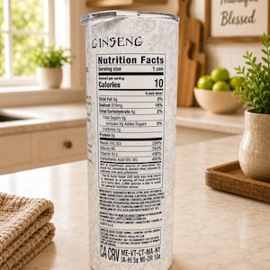 K&ouml;nnte beinhalten: Ein zylindrischer wei&szlig;er Becher mit einem schwarz aufgedruckten N&auml;hrwertetikett und dem Wort GINSENG am oberen Rand. Das Design enth&auml;lt detaillierte Angaben zu Portionsgr&ouml;&szlig;e, Kalorien, Vitaminen und Inhaltsstoffen sowie einen transparenten Deckel.