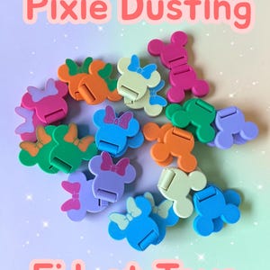 May include: A collection of colorful plastic fidget toys shaped like mouse heads with bows, featuring various solid colors including pink, orange, green, blue, and purple. The text Pixie Dusting Fidget Toys is displayed in large pink letters.