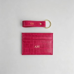 May include: A fuchsia-colored leather keychain and cardholder set. The keychain has a gold-colored ring and the cardholder has multiple slots. Both items are embossed with a crocodile pattern and the initials "AM" in gold lettering.