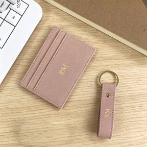 Peut inclure: Un porte-cartes rose blush et un porte-clés assorti, tous deux avec les initiales dorées « RM », sont présentés sur une surface en bois clair. Le porte-cartes comporte plusieurs fentes et le porte-clés est doté d'un anneau doré. Accessoires personnalisés.