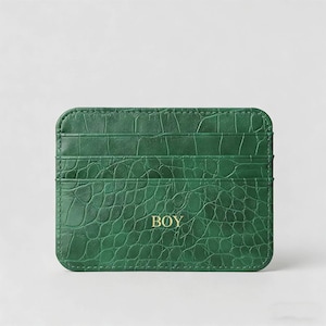Puede incluir: Tarjetero verde con textura de piel de cocodrilo. El tarjetero tiene múltiples ranuras y la palabra "BOY" en letras doradas. El tarjetero es rectangular con esquinas redondeadas.