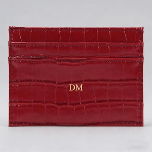 May include: A red card holder with a textured, crocodile-effect pattern. The card holder has multiple slots and the initials "DM" in gold lettering. The design is sleek and compact.