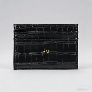 May include: Black card holder with a crocodile texture. The card holder has multiple slots and the gold letters "AM" are visible on the front. The card holder is made of a shiny material.