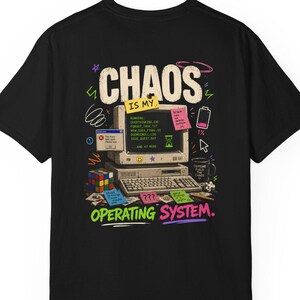 May include: A black t-shirt featuring a graphic of a vintage computer with the text "CHAOS IS MY OPERATING SYSTEM." The design includes colourful sticky notes, a Rubik's cube, a coffee mug, and a low battery icon, all arranged around the computer.