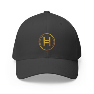 May include: A charcoal grey baseball cap featuring a centred, circular gold embroidered emblem. The emblem consists of a gold circle outline surrounding a stylised capital letter H with a horizontal line through the middle, rendered in gold.
