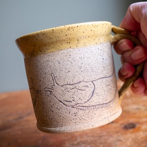May include: Handmade ceramic mug with a tan and speckled finish, featuring a dark brown etched design of a snail. The rim and handle are a solid tan color. The mug is held by a person's hand.