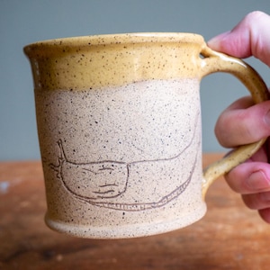 May include: A handmade ceramic mug with a tan base and a yellow rim. The mug features a hand-drawn illustration of a slug. The mug is held by a person's hand.