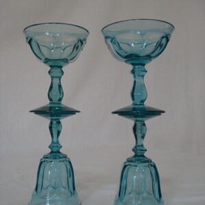 May include: Two vintage teal-colored champagne coupe glasses. Each glass features a rounded bowl, a slender stem, and a flared base. The glasses are made of clear glass and have a delicate, ornate design. The glasses are displayed against a plain white background.