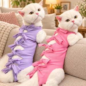 May include: Two white cats wearing ribbed sleeveless shirts. One cat wears a light purple shirt with a vertical row of five tied bows down the front. The other cat wears a bright pink shirt with a matching vertical row of five tied bows down the front.