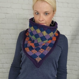 Puede incluir: Una mujer lleva un calentador de cuello de ganchillo a cuadros en morado, naranja y verde. El calentador de cuello tiene forma triangular y tiene un borde de ganchillo.