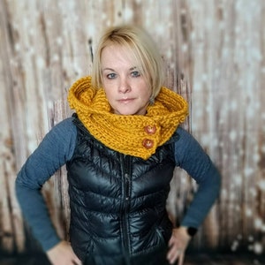 May include: A yellow knitted cowl neck scarf with two wooden buttons. The scarf is worn by a person with blonde hair and is styled to cover the neck and ears.