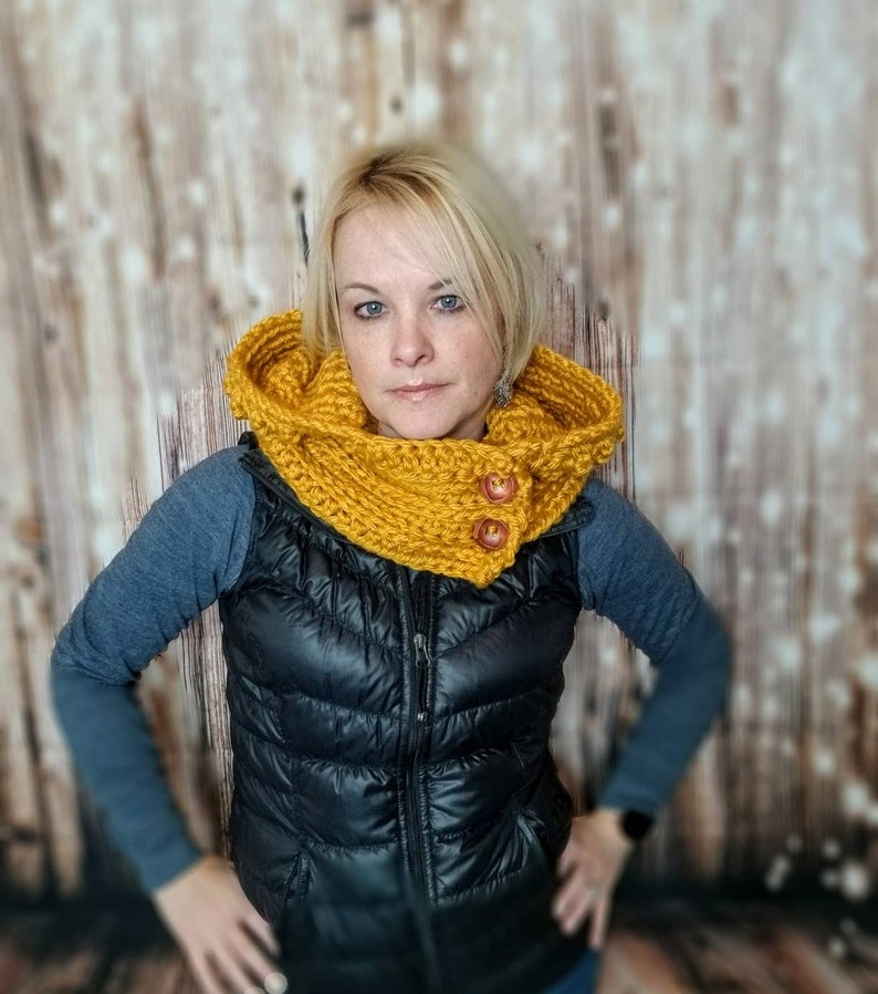 May include: A yellow knitted cowl neck scarf with two wooden buttons. The scarf is worn by a person with blonde hair and is styled to cover the neck and ears.