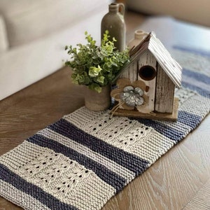 May include: A knitted table runner with a white and blue striped pattern. The runner is decorated with a small wooden birdhouse and a potted plant.