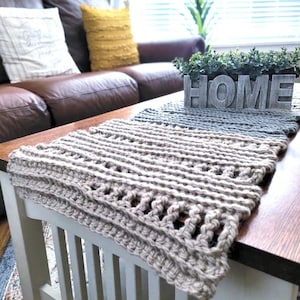 Könnte beinhalten: Beige handgestrickter Tischläufer mit strukturiertem Muster, platziert auf einem Couchtisch aus dunklem Holz. Ein HOME-Schild aus Holz und ein kleiner Pflanzkasten befinden sich ebenfalls auf dem Tisch. Im Hintergrund ist ein braunes Ledersofa zu sehen.