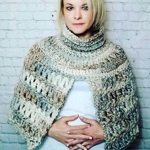 May include: A hand-crocheted shawl in cream, grey, and brown. The shawl has a high, ribbed collar and a textured, open weave design. The garment is worn over a white top.