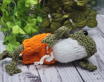 May include: A knitted frog wearing orange shorts. The frog is lying on its side with its legs outstretched. The frog is made of green, white, and orange yarn.