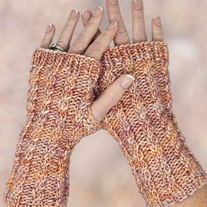 Könnte beinhalten: Ein Paar handgestrickte Fingerhandschuhe in warmen Farben wie Orange, Pfirsich und Creme. Die Handschuhe haben ein strukturiertes Zopfmuster und sind aus dickem Garn gefertigt, ideal um die Hände warm zu halten.