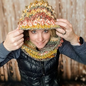 May include: A multi-colored crocheted hooded scarf with a textured pattern. The scarf is made with shades of green, brown, orange, and white yarn.