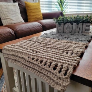 May include: A hand-crocheted table runner in shades of beige, grey, and tan. The runner is placed on a dark wood coffee table. A wooden sign that says "HOME" is on the table. Decorative cushions are on a brown leather sofa in the background.