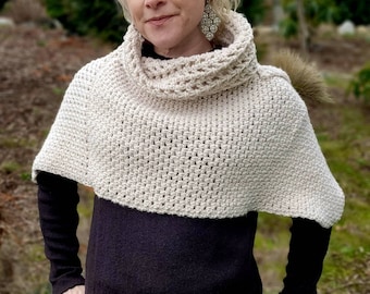May include: A cream-colored crochet cowl neck poncho featuring a textured waffle stitch pattern. The garment has a wide, folded collar and a short, cape-like silhouette that drapes over the shoulders, worn over a black long-sleeve shirt.