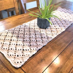 May include: A cream, tan, and grey crochet table runner. The openwork design is placed on a wooden table, with a small potted plant on top. The runner adds a touch of rustic charm to any dining space. The table runner is approximately 150 cm long.