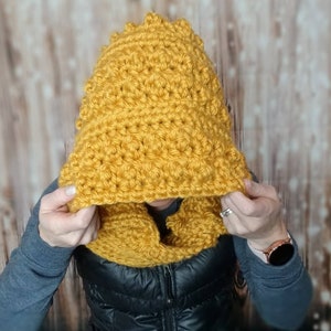 May include: A yellow crocheted hooded scarf with a textured stitch pattern. The scarf is worn by a person, but the person's face is not visible.