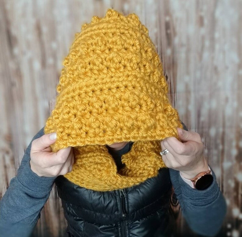May include: A yellow crocheted hooded scarf with a textured stitch pattern. The scarf is worn by a person, but the person's face is not visible.