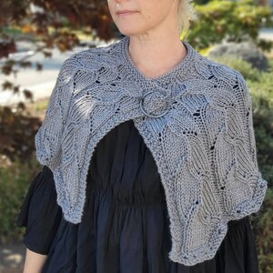 May include: A grey knitted shawl with a leaf pattern and a circular metal clasp. The shawl is draped over the shoulders. The garment is made of a soft, textured yarn. The background is blurred, with green and brown tones.