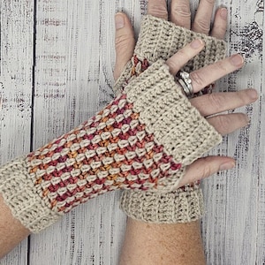 May include: A pair of beige crochet fingerless gloves. The gloves have a textured pattern in shades of red, orange, and yellow. The cuffs and upper portion of the gloves are a solid beige color. The gloves are displayed on a light wood surface.