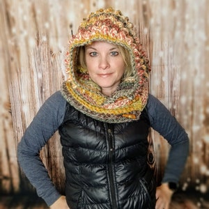 May include: A woman wearing a crocheted hooded scarf in shades of orange, yellow, and green. The scarf is made with a textured stitch and has a unique, almost spiky, design. She is also wearing a black puffer vest.