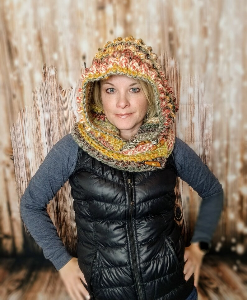 May include: A woman wearing a crocheted hooded scarf in shades of orange, yellow, and green. The scarf is made with a textured stitch and has a unique, almost spiky, design. She is also wearing a black puffer vest.