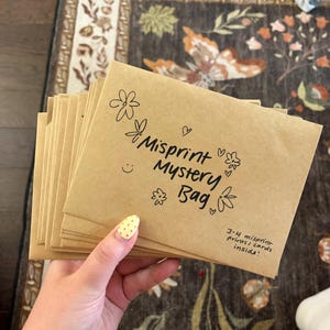 May include: A stack of light brown kraft paper envelopes featuring black hand-drawn floral doodles, hearts, a smiley face, and the text "Misprint Mystery Bag" with "3-4 misprint prints/cards inside!" written in the bottom right corner.