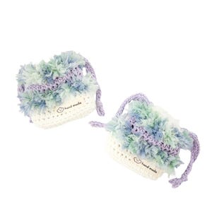 Puede incluir: Dos pequeñas bolsas con cordón hechas de ganchillo blanco en la base con una sección superior texturizada y peluda en tonos azul claro, verde y lavanda. Cada bolsa presenta una pequeña etiqueta de tela blanca con el texto hand made y un icono de nube.