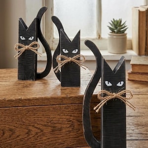 May include: Three black wooden cat figurines with pointed ears, white painted eyes, and a natural jute twine bow tied around each neck. Each cat has a curved tail extending from the side, and the wood features a distressed, dark finish.