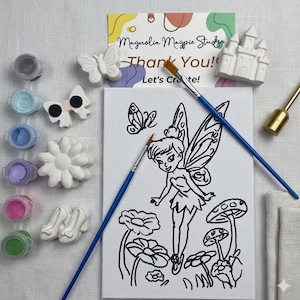 May include: A paint-your-own fairy-themed craft kit. The kit includes a canvas with a fairy outline, paint pots, paintbrushes, and various ceramic shapes like a butterfly, bow, flower, and castle. The text on the card reads "Magnolia Magpie Studio - Thank You! Let's Create!"