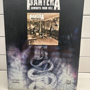 May include: A Pantera Cowboys From Hell authentic guitar tab edition music book. The black cover features the band name, album title, a central photograph of the band in a room, a large coiled snake graphic, a Parental Advisory label, and Alfred logo.